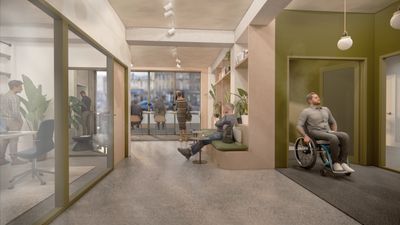 Inside of an office building with a man in a wheelchair coming out of a lift and people at hot desks