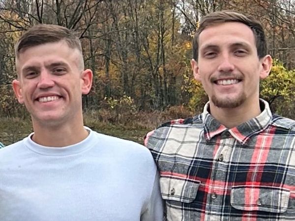 Two smiling young men posing outdoors with autumn trees in the background.