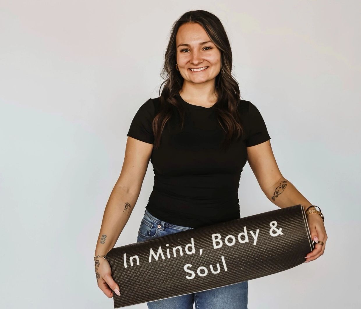 A smiling woman holding a rolled yoga mat with the words 'In Mind, Body & Soul'.