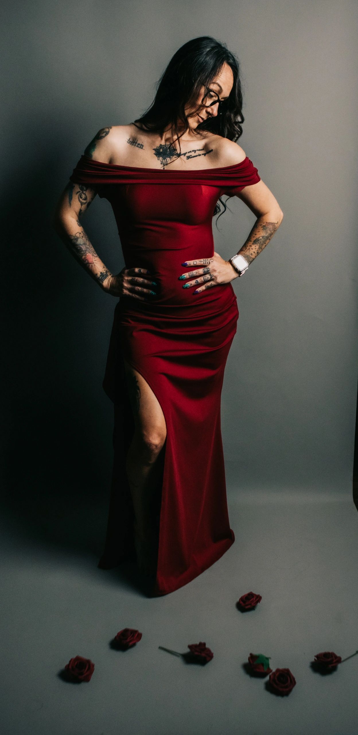 Woman in a red gown with tattoos and glasses, standing among scattered roses.