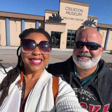 A smiling couple wearing sunglasses poses in front of Creation Museum Welcome Center.