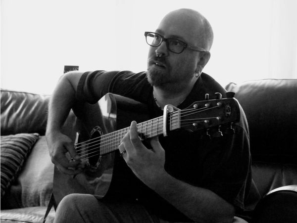 Man with glasses playing an acoustic guitar on a couch.