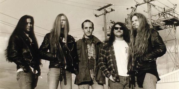 Five men with long hair and leather jackets standing outdoors under power lines.