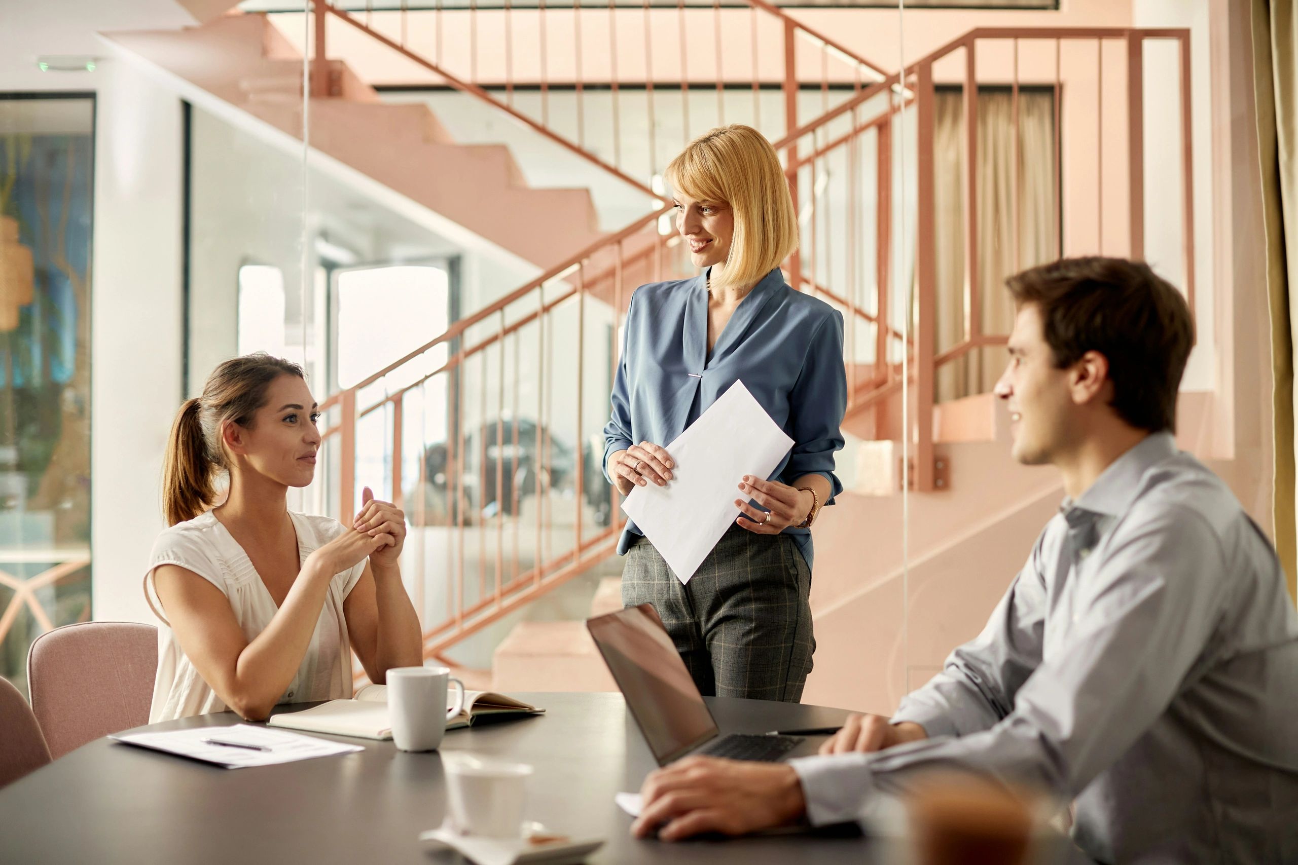 Three colleagues engaged in a friendly office discussion near a staircase.