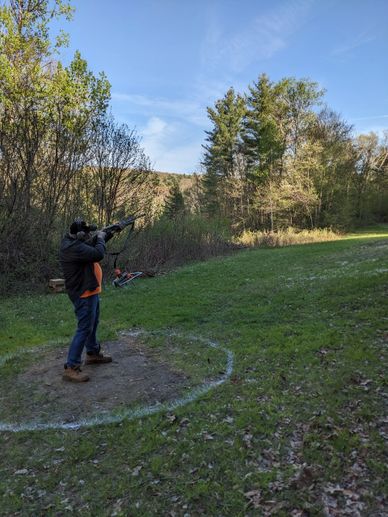 Person aiming a rifle outdoors in a wooded area.