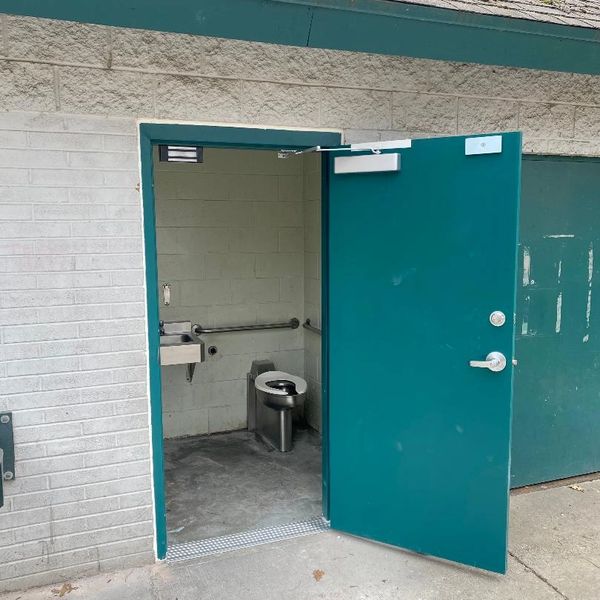 Outdoor public restroom with an open teal door revealing a stainless steel toilet and sink.