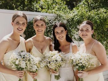 Four women in elegant dresses holding bouquets, smiling outdoors.