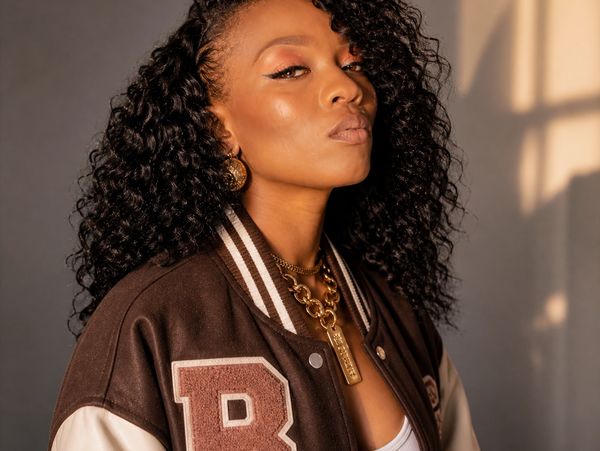 Confident woman with curly hair in a brown varsity jacket and gold jewelry.