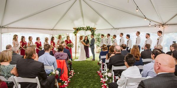 A wedding ceremony with bride and groom under a decorated arch inside a tent.