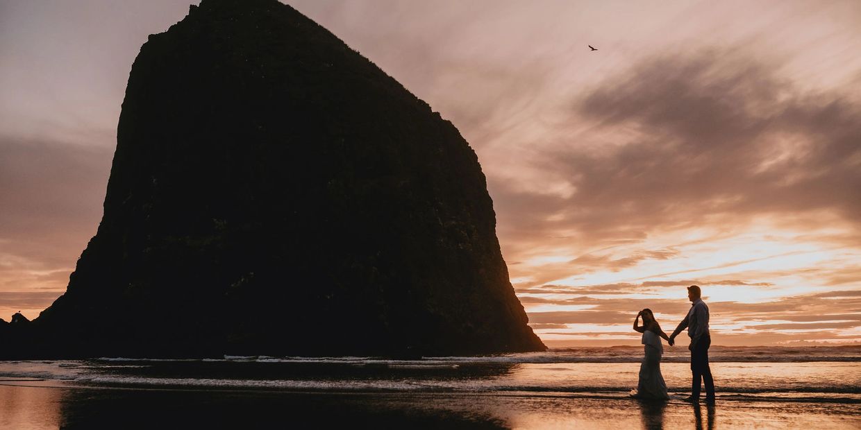 A couple holding hands walks along the beach at sunset with a large rock formation.