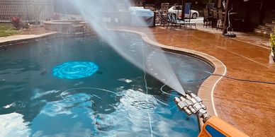 Hand holding an orange water spray gun over a backyard pool on a sunny day.