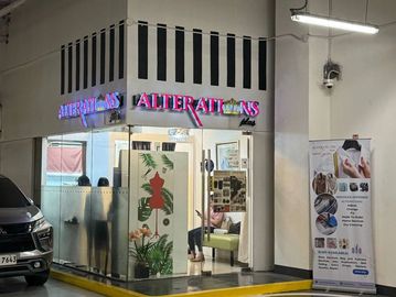 A corner alteration shop with glass walls in a parking area, featuring a neon sign and a waiting area inside.