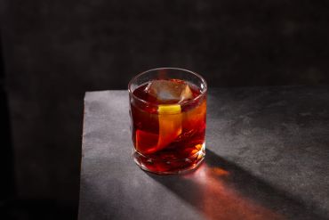 A glass of red cocktail with a lemon peel on a dark surface.