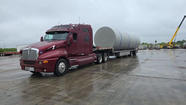 Red semi truck hauling a large cylindrical tank on a wet concrete surface.