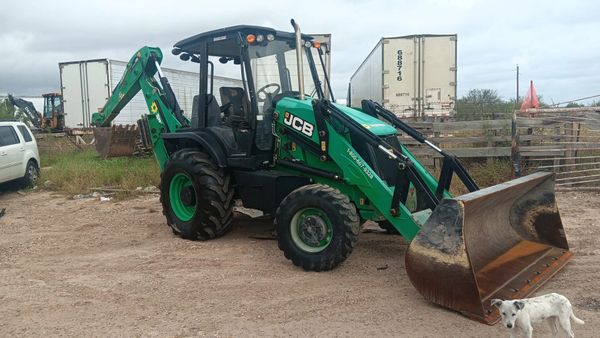 A green JCB backhoe loader parked on a dirt ground with a small white dog nearby.