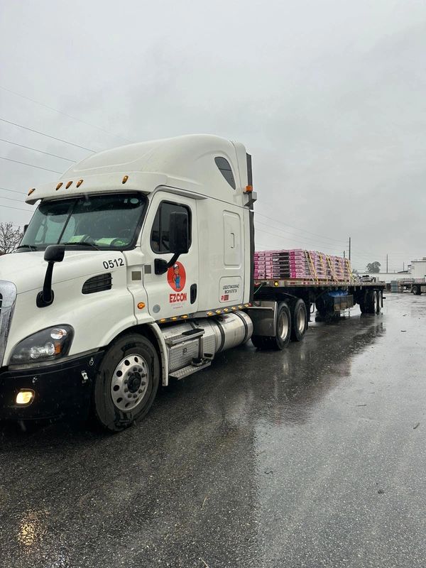 White semi-truck with flatbed trailer loaded with palletized goods on wet pavement.