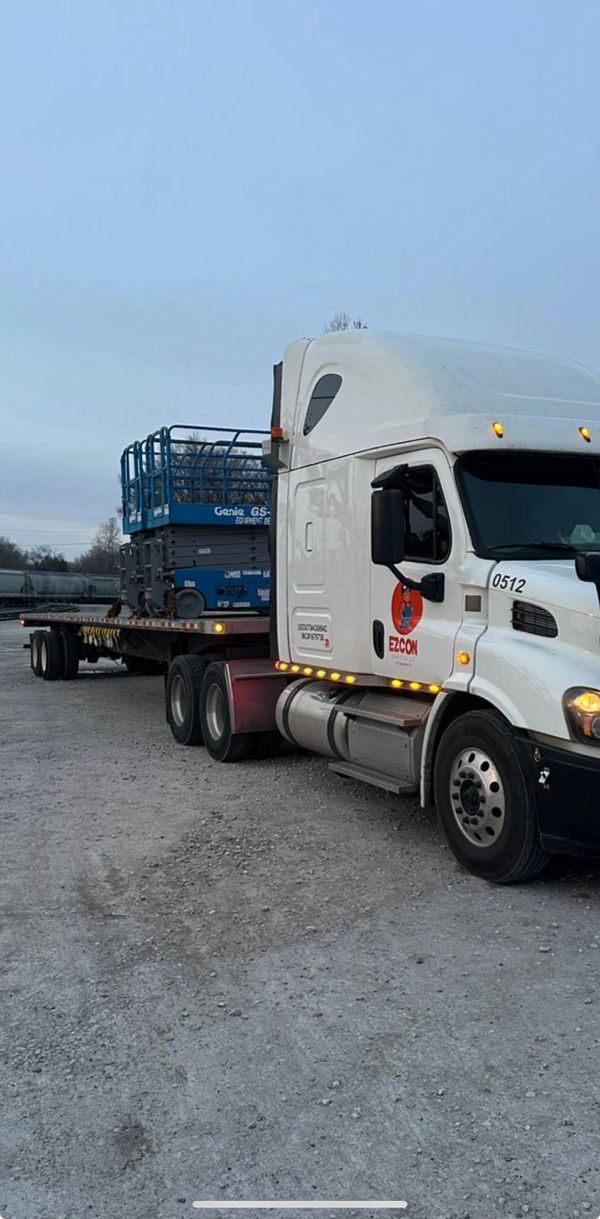 White semi-truck with a flatbed trailer carrying a blue Genie scissor lift.