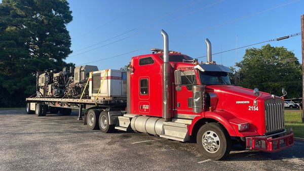 Red Kenworth semi truck parked with a flatbed trailer carrying heavy equipment.