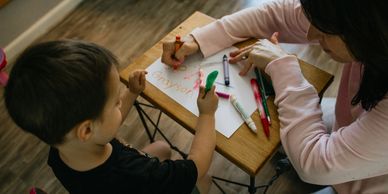 A dark haired teacher coloring a page and teaching a toddler how to write letters.