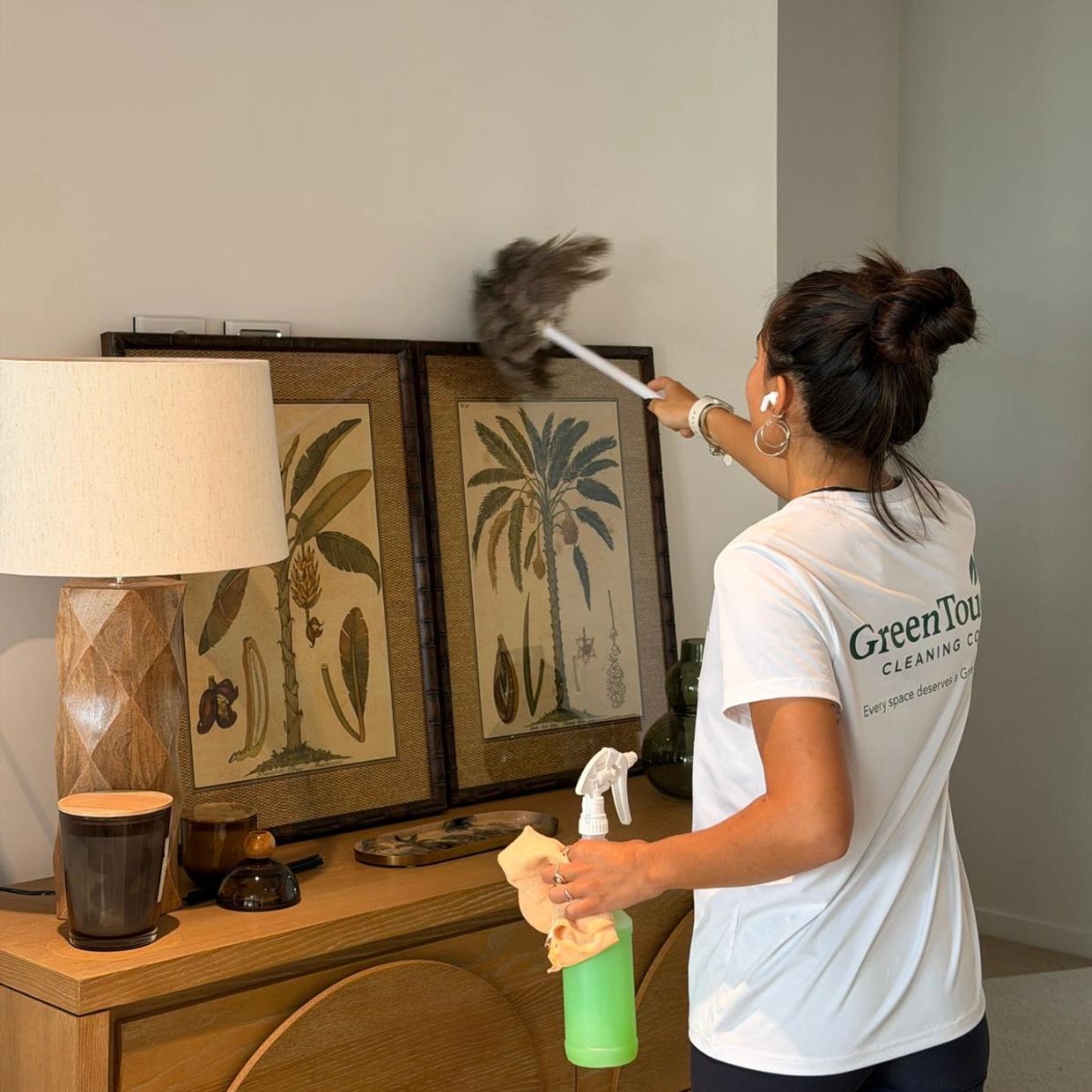 Woman dusting framed artwork on a wooden sideboard.