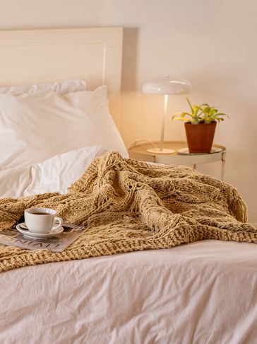 Cozy bed with a knitted blanket, cup of coffee, and a magazine in a softly lit room.