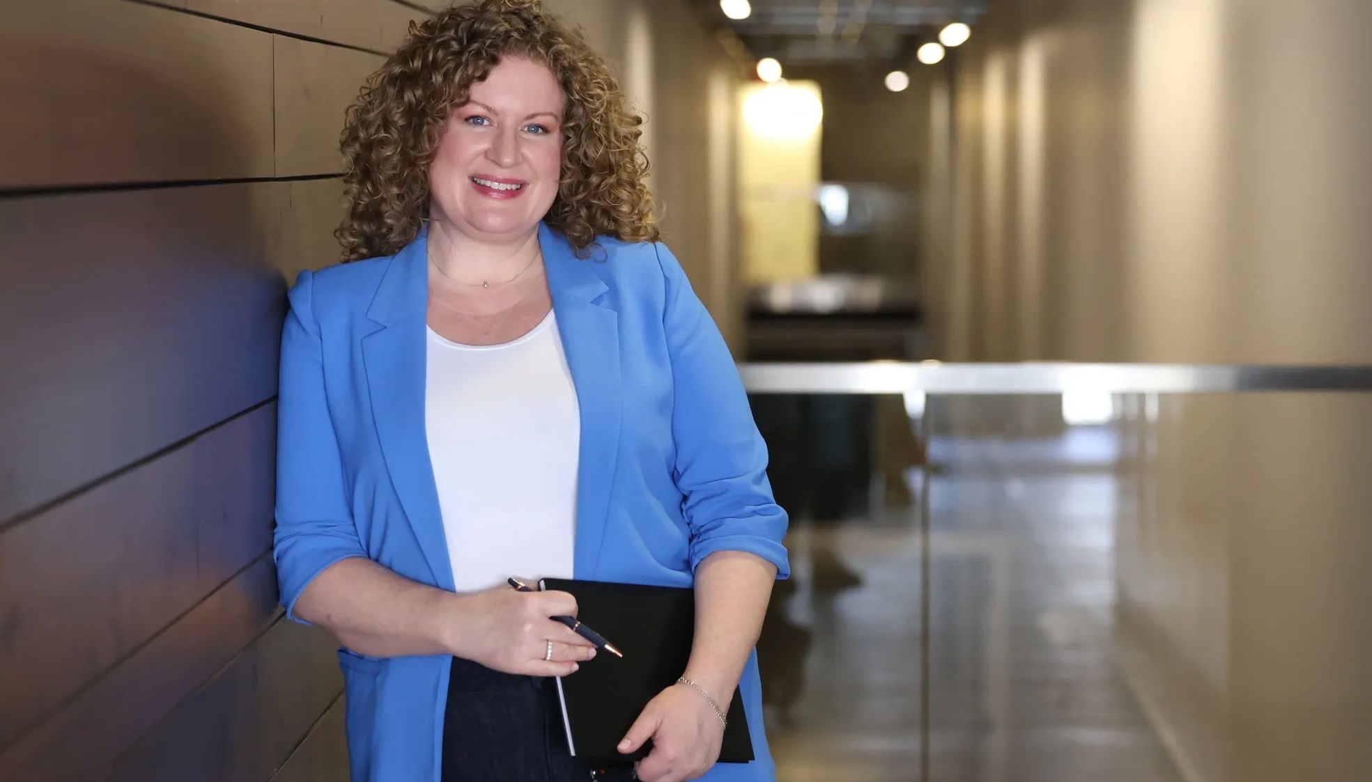 Woman with curly hair wearing a blue jacket, holding and book and pen, is leaning against a wall. 