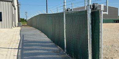 A long green chain-link fence with barbed wire on top beside a concrete path.