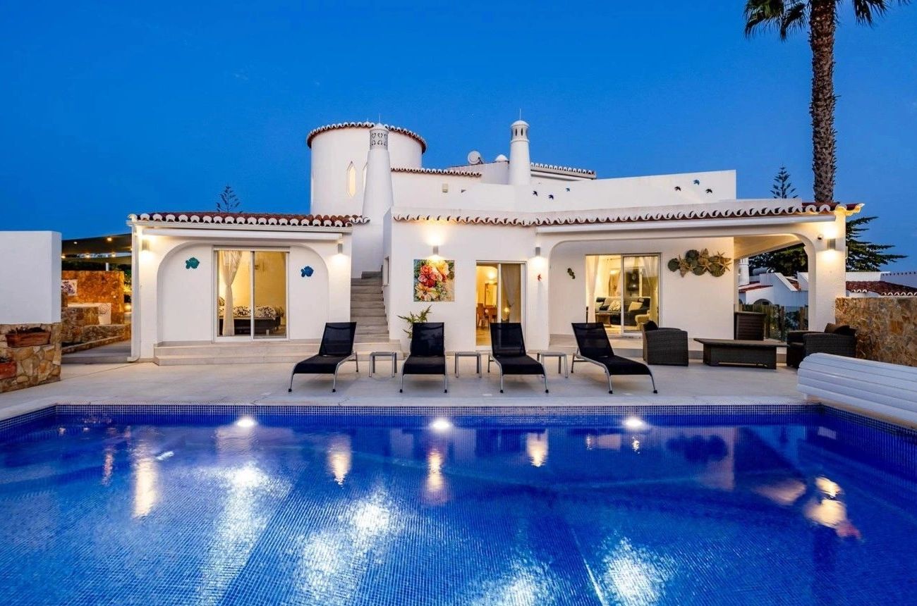 Modern white villa with a lit pool and lounge chairs at dusk.