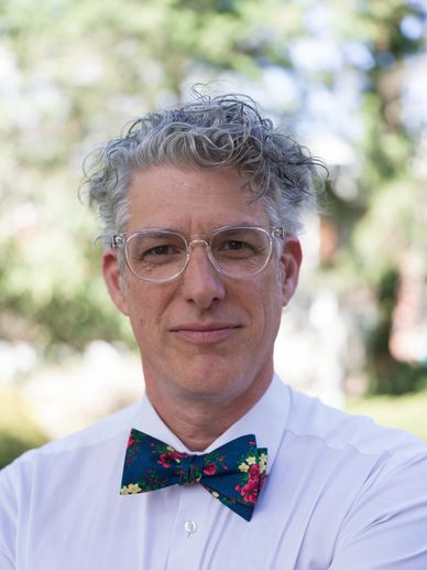 Man with curly gray hair, glasses, and a floral bow tie smiling outdoors.