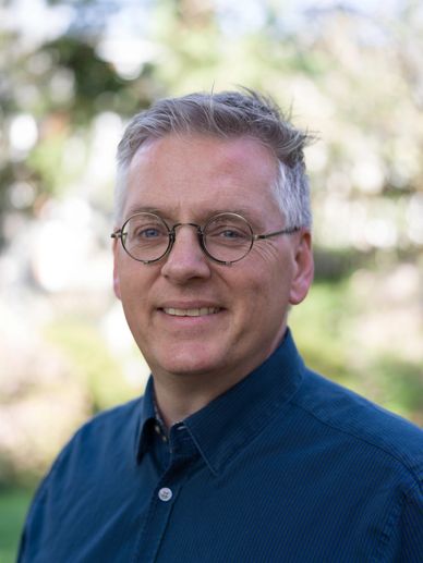 Smiling man with glasses wearing a blue shirt outdoors.