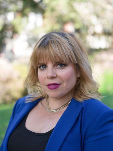 Woman with blonde hair wearing a blue blazer and purple lipstick outdoors.