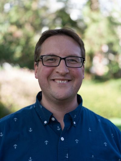 Smiling man wearing glasses and a navy blue anchor-patterned shirt outdoors.
