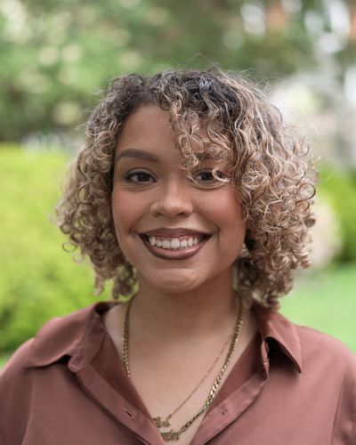 Smiling woman with curly hair outdoors in a brown shirt.