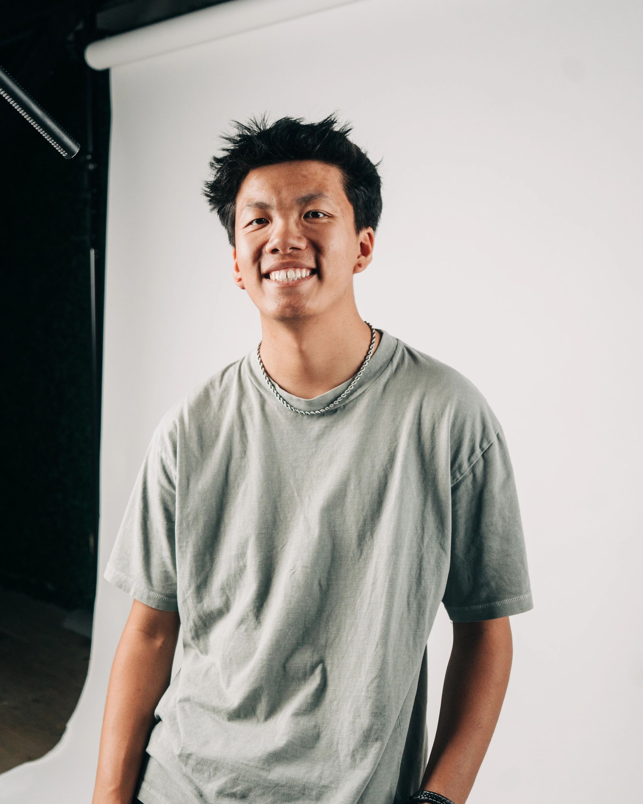 Young man smiling in a casual t-shirt with a necklace against a plain backdrop.