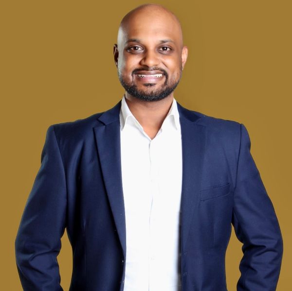Smiling man in a navy blazer and white shirt against a solid tan background.