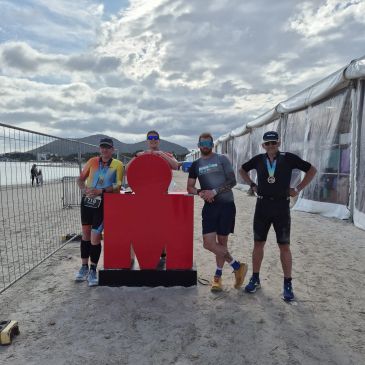 Four athletes posing beside a large red Ironman symbol on a beach.