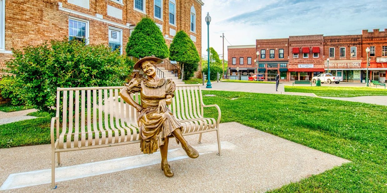 Bronze statue of a woman sitting on a bench in a small town square.