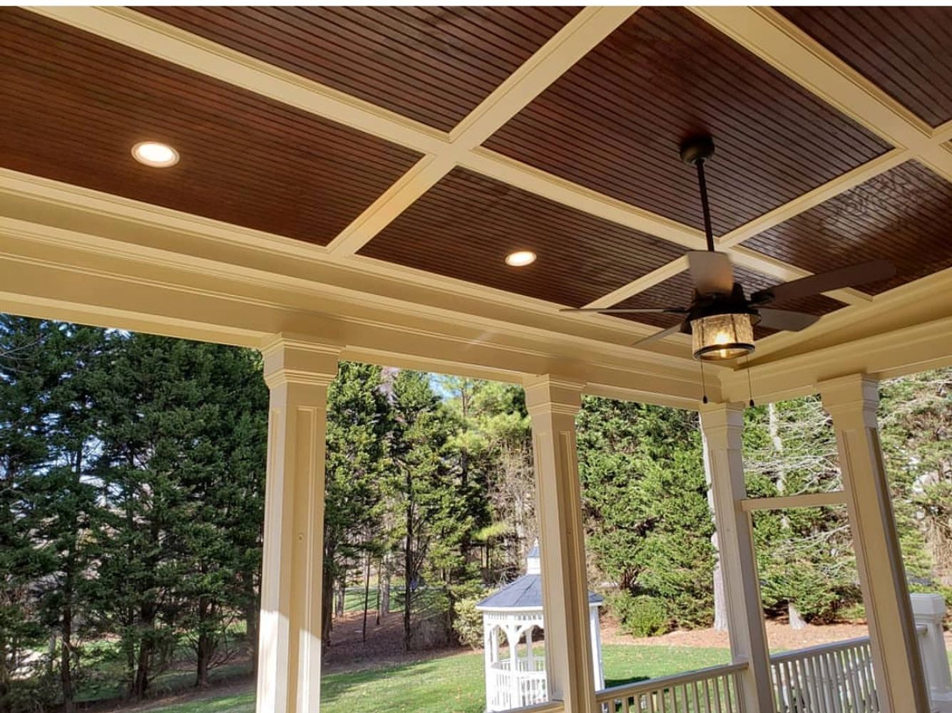 Porch with wooden ceiling, fan, and view of green trees and gazebo.
