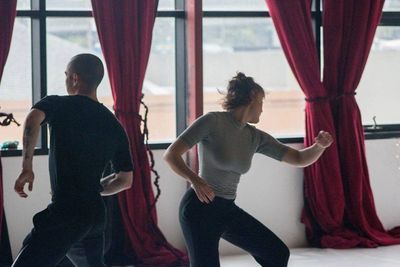 Two dancers moving away from each other, in front of bay windows that look out on a cityscape.