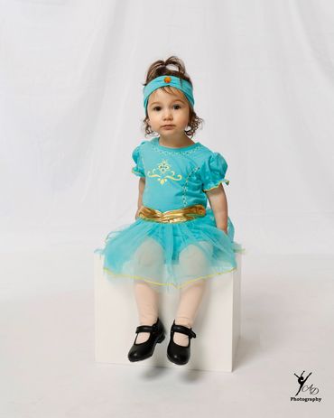 A little girl in a turquoise princess dress sitting on a white cube.