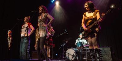 Female band performing on stage with instruments and microphones.