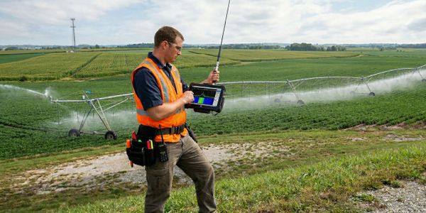 Agricultural worker monitors irrigation system with tablet and radio in vast green field.