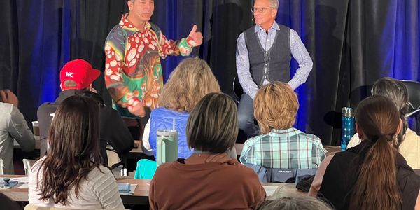 Two men speaking on stage to an audience seated at tables.