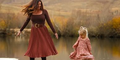 A woman and young girl joyfully twirling near a calm lake in autumn.