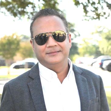 Man in sunglasses and blazer smiling outdoors on a sunny day.