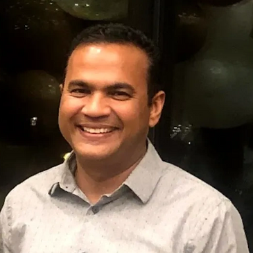 Smiling man in a light grey shirt, posing indoors with a warm expression.