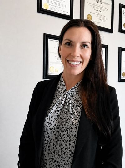 Professional woman smiling with framed certificates on the wall.