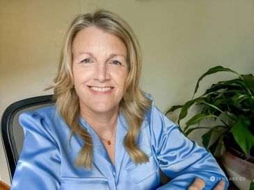Smiling woman in a blue satin blouse sitting indoors with a plant in the background.