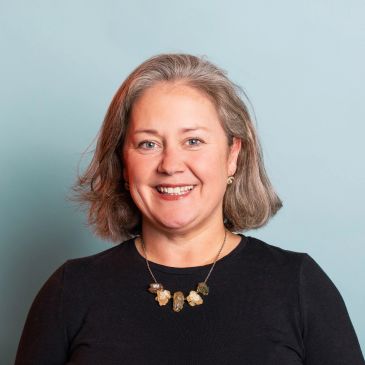 Smiling woman with gray hair and a statement necklace against a light blue background.