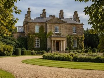 A stately stone mansion surrounded by lush gardens and a curved gravel driveway.